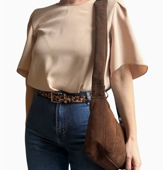 woman wearing a beige blouse, blue jeans, and holding a brown suede bag on a plain background