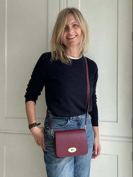 Woman wearing a black sweater and blue jeans with a burgundy handbag, standing in a room with white walls.