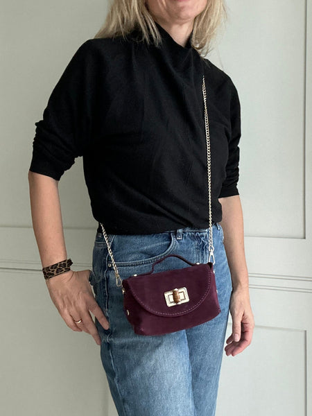 Woman wearing a black top and blue jeans with a maroon handbag, standing in front of a white door.