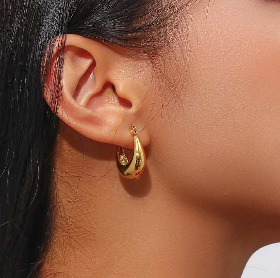 A close-up of a person's ear wearing a polished gold huggie hoop earring, with dark hair and warm sunlight highlighting the jewelry.
