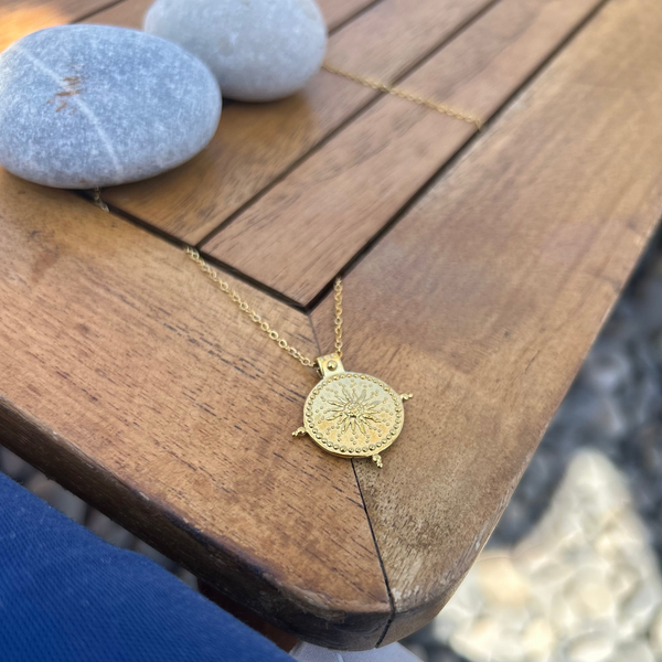 A gold pendant necklace with an engraved sunburst design on a delicate chain, resting on a wooden table next to smooth gray stones.
