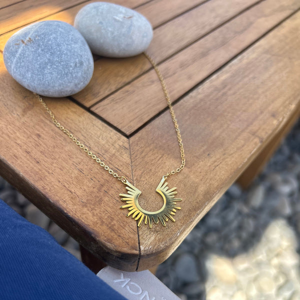 A gold necklace with sunburst rays is displayed on a wooden table outdoors, next to two smooth gray stones. 