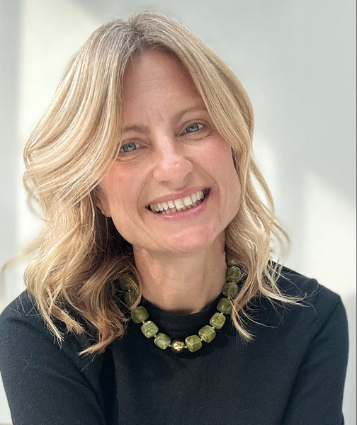 Woman with blonde hair wearing a black top and green necklace against a white background