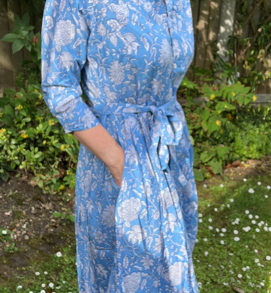 woman with hands in pockets of a blue floral dress standing in a garden.