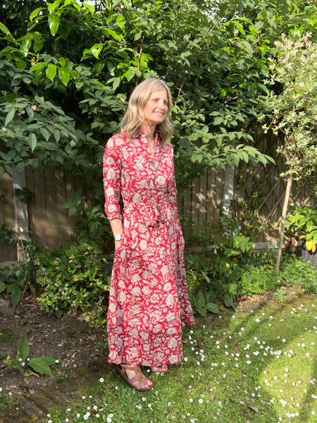 Woman in a red floral dress standing in a garden with greenery and flowers.