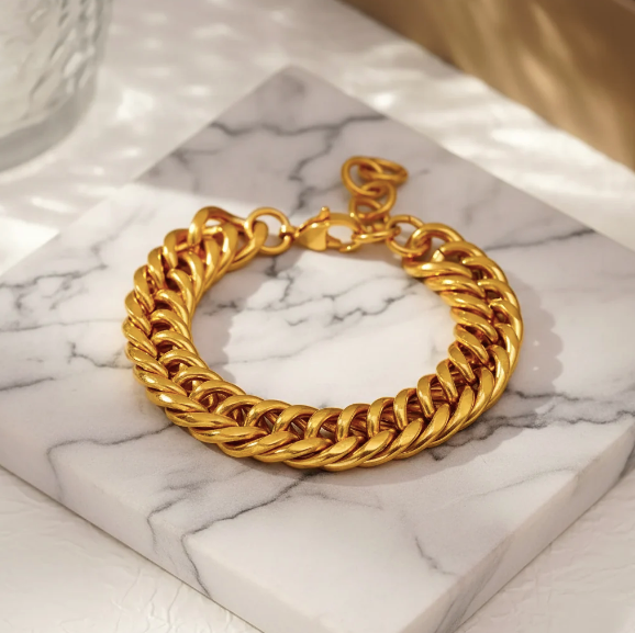 A chunky gold-tone Cuban link chain bracelet resting on a white marble block with gray veining, set against a softly lit neutral background.