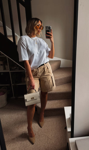 A woman takes a mirror selfie in a home setting, wearing a white short-sleeve t-shirt, beige cargo shorts, beige pointed-toe heels, and holding a small white handbag.