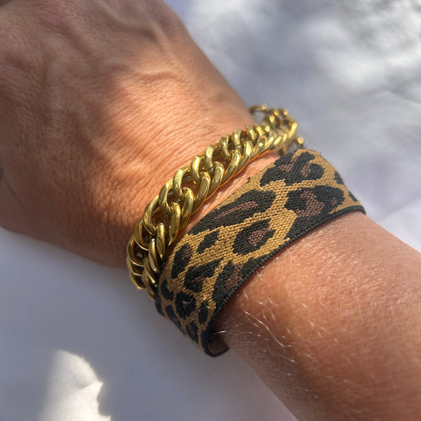 A close-up of a person's wrist wearing two bracelets: a thick gold curb chain and a wide black and tan leopard print fabric cuff bracelet, set against a white fabric background.