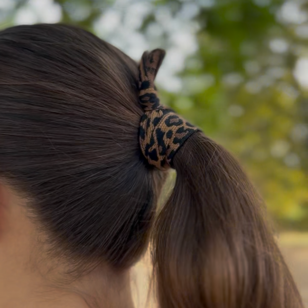 A close-up shot of a person from behind, showing a brown ponytail secured with a leopard-print fabric scrunchie outdoors with a blurred green background.