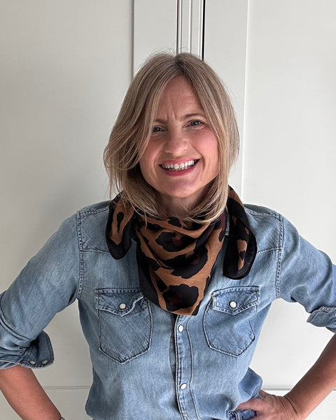 A smiling blonde woman posing indoors against white paneled doors, wearing a light blue denim shirt and jeans with a brown and black leopard-print neck warmer scarf.