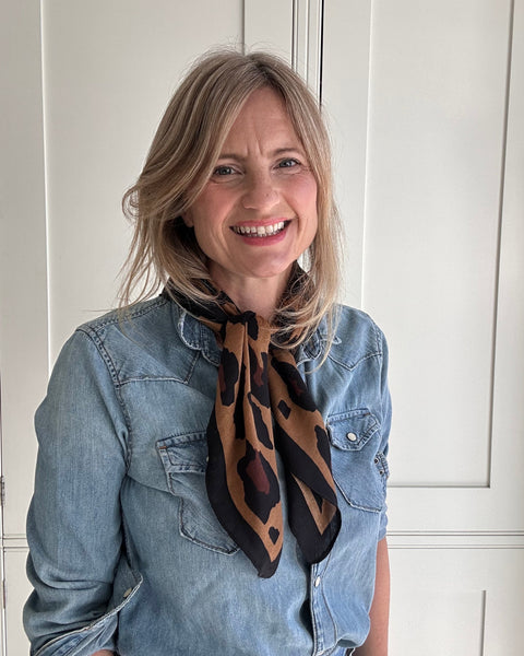 A smiling blonde woman wearing a light blue denim shirt and a brown and black patterned scarf, standing in front of white paneled doors.