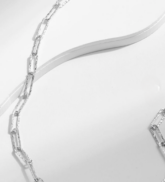 A close-up of a silver-toned chain necklace with elongated oval links, draped diagonally across a white surface.