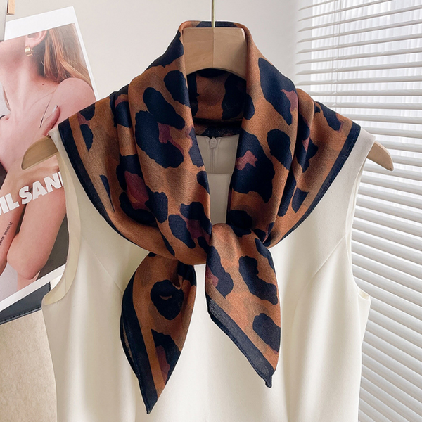A leopard print scarf draped over a cream-colored sleeveless top on a wooden hanger, with a magazine cover partially visible on the left and white window blinds in the background.