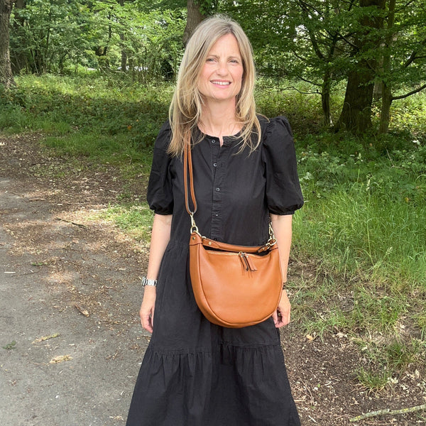 A woman standing outdoors on a dirt path in a wooded area, wearing a black midi dress and black sandals while carrying a tan leather crescent-shaped crossbody bag.
