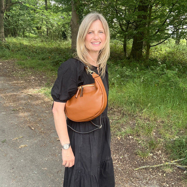 A woman with blonde hair stands outdoors on a dirt path in a wooded area, wearing a black dress and carrying a brown leather crescent-shaped shoulder bag.