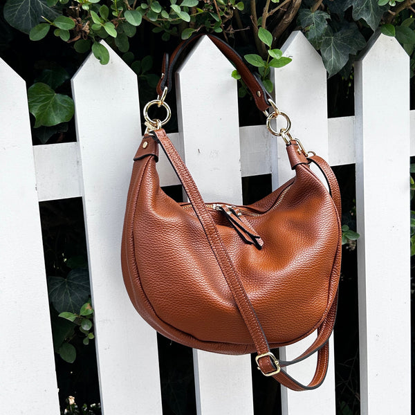 A brown pebbled leather crescent-shaped shoulder bag with a top handle and adjustable crossbody strap, hanging in front of a white picket fence with green ivy in the background.
