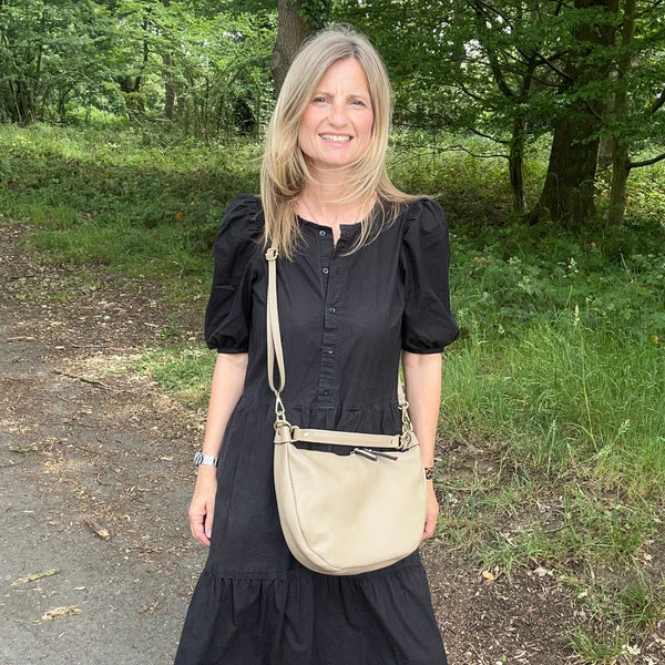 A woman standing outdoors on a gravel path in a wooded area, wearing a black midi dress with puff sleeves, a beige crossbody bag, and black sandals.