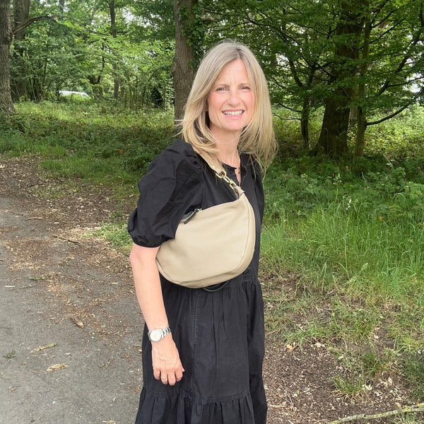A woman with blonde hair stands outdoors on a path wearing a black midi dress, a beige shoulder bag, and a gold necklace, with trees and grass in the background.