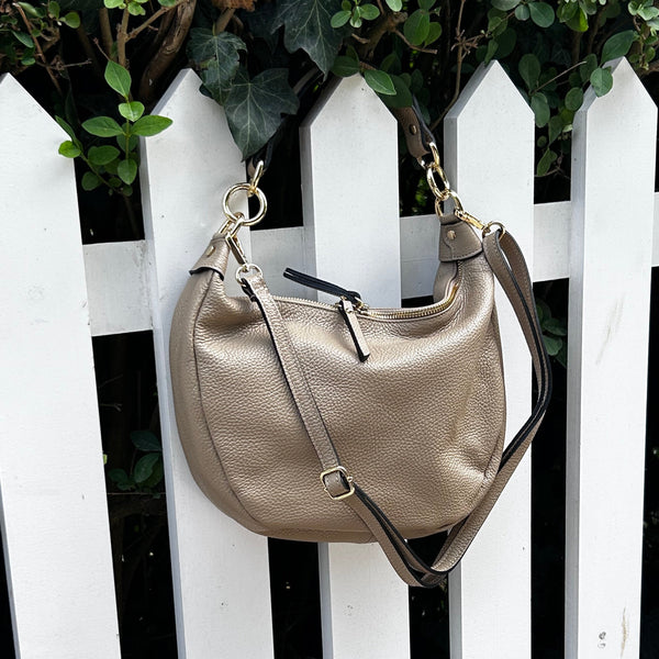 A taupe pebbled leather shoulder bag with gold-tone hardware, hanging from a white picket fence with green ivy in the background.