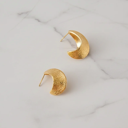 A pair of textured gold crescent-shaped hoop earrings resting on a white marble surface.