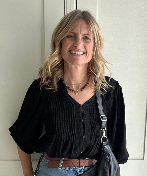 Woman wearing a black top and blue jeans with a black bag and gold jewellery, standing against a white wall.