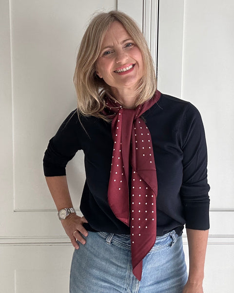 A smiling blonde woman wearing a black long-sleeve top, light blue jeans, and a burgundy silk scarf with small white polka dots, standing in front of white paneled doors.
