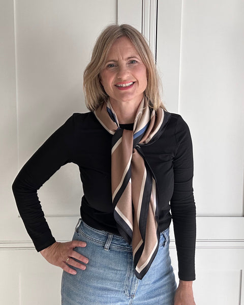 A woman with blonde hair wearing a black long-sleeve top, light blue jeans, and a beige and black striped silk scarf, standing in front of white paneled doors.