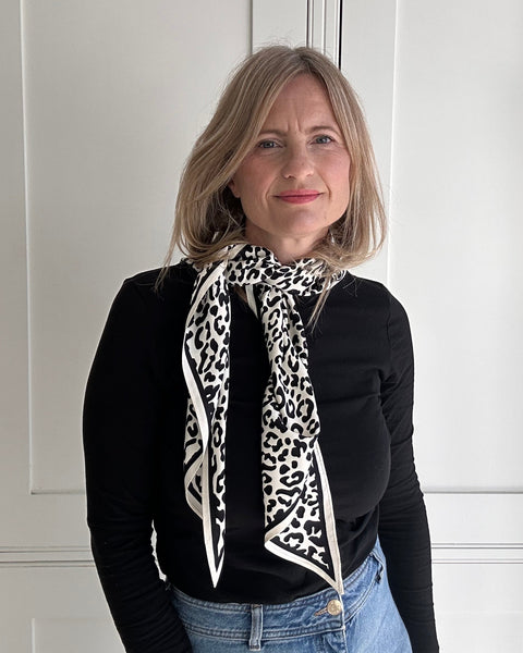 A woman with blonde hair wearing a black long-sleeve top, blue jeans, and a black and white leopard print scarf tied around her neck, standing in front of white paneled doors.