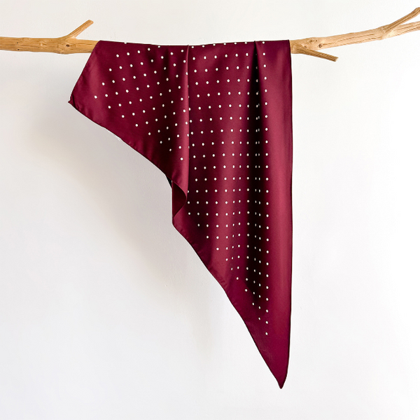 A deep burgundy triangle scarf with a white polka-dot pattern, draped over a light brown wooden branch against a plain white background.