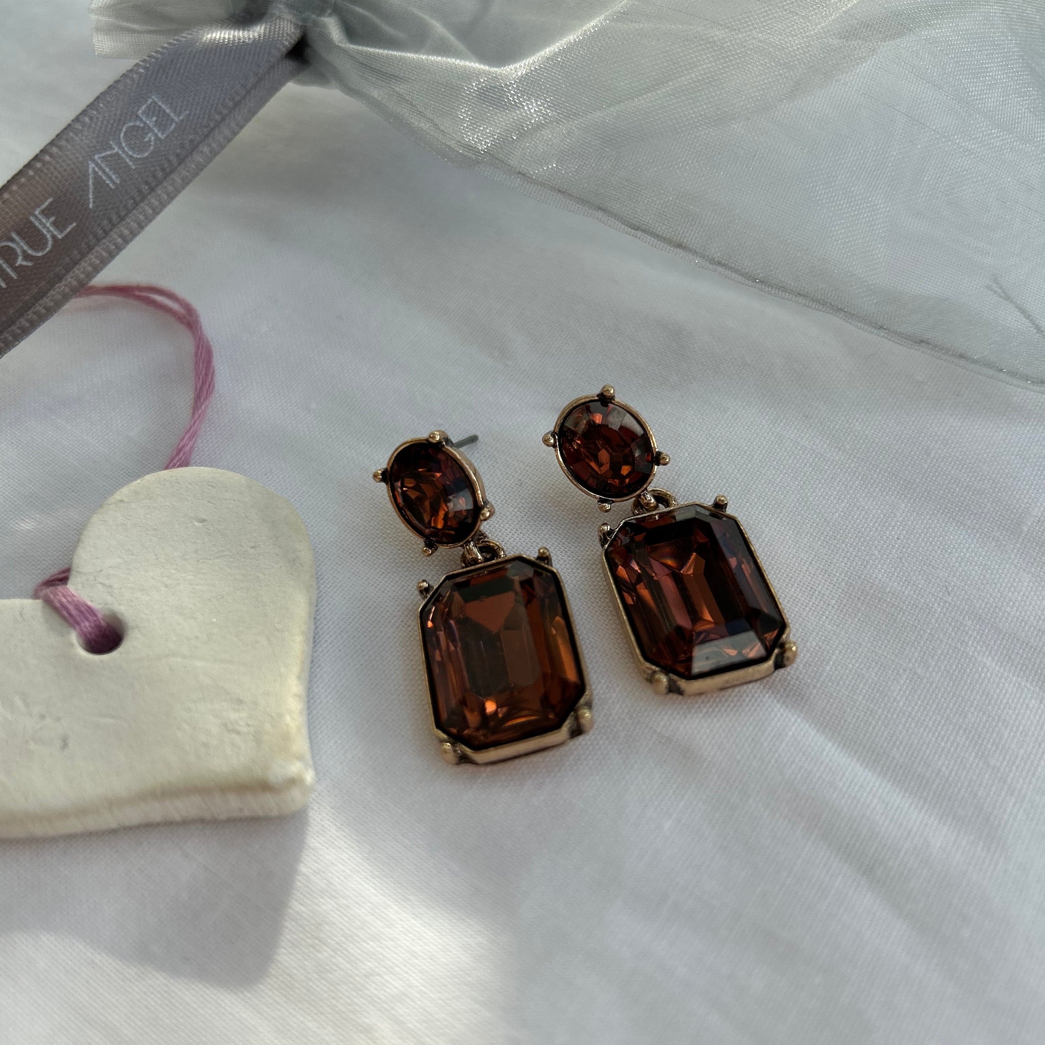 Pair of brown gemstone earrings on a white fabric background with a heart-shaped object and 'Last True  Angel' ribbon.