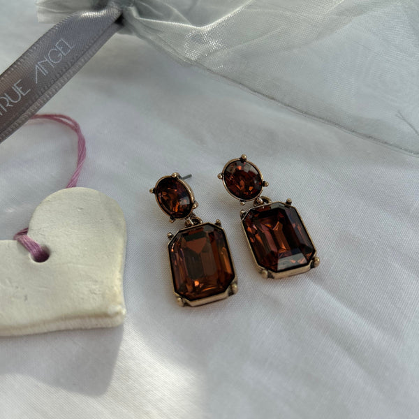 Pair of brown gemstone earrings on a white fabric background with a heart-shaped object and 'Last True  Angel' ribbon.