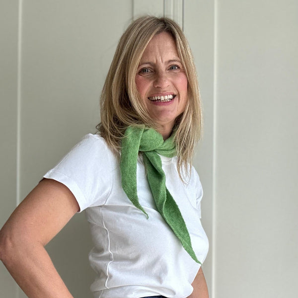 Woman wearing a white shirt, blue jeans, and a green scarf standing against a white wall.