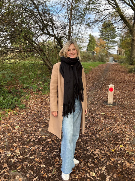 Woman standing on a leaf-covered path wearing a beige coat, black scarf, and light blue jeans.