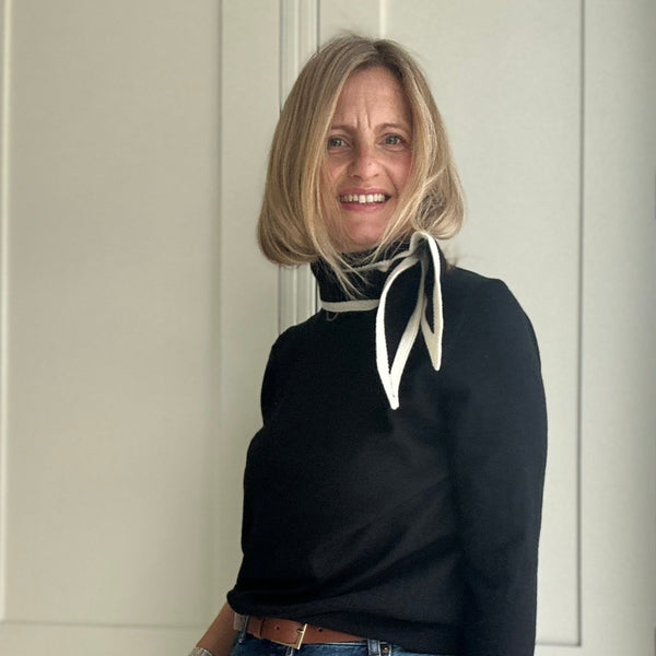 Woman in black sweater with a black sand cream tied scarf against a pale wood background