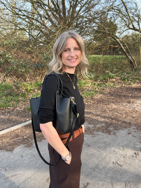 woman in black sweater and brown trousers carrying a black leatehr handbag standing in a forest