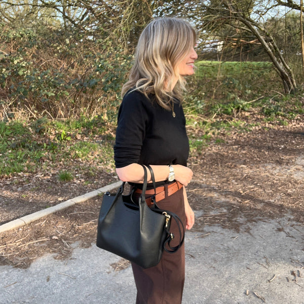 woman in black sweater and brown trousers carrying a black leatehr handbag standing in a forest