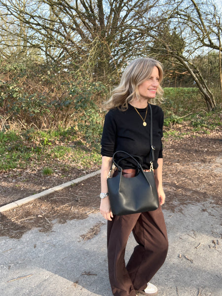 woman in black sweater and brown trousers carrying a black leatehr handbag standing in a forest