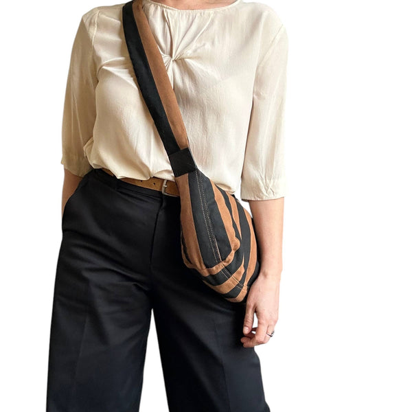 A woman wearing a cream blouse and black trousers carries a brown and black striped crossbody sling bag against her side.