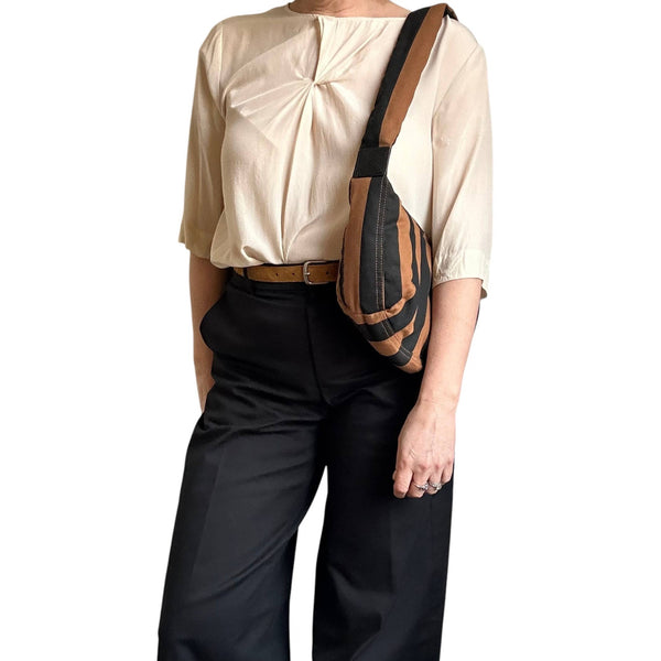 A person wearing a cream pleated blouse, black trousers, and a brown belt, carrying a brown and black striped crossbody bag.