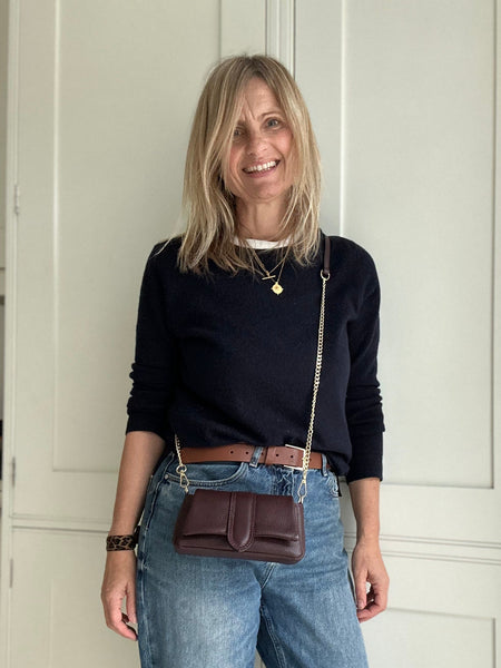 Woman wearing a black top, blue jeans, and carrying a burgundy leather bag in front of a white door.