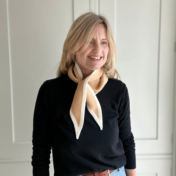 Woman wearing a black sweater and blue jeans with a beige scarf, standing against a white paneled wall.