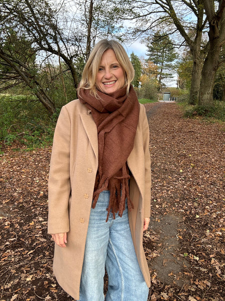 Woman wearing a beige coat and brown scarf standing on a leaf-covered path in a park.
