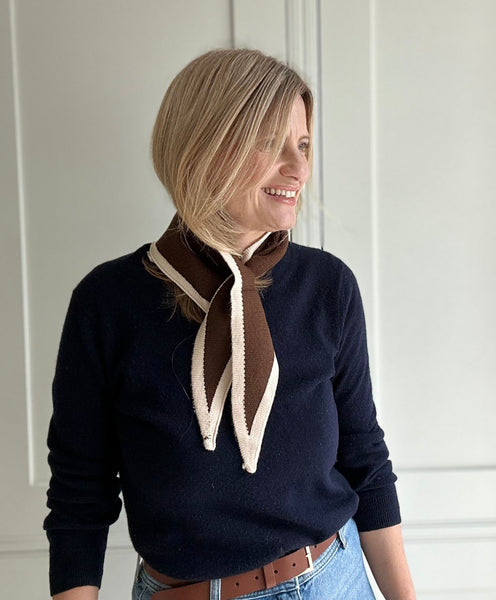 Woman wearing a navy sweater with a brown and white scarf against a white paneled wall.
