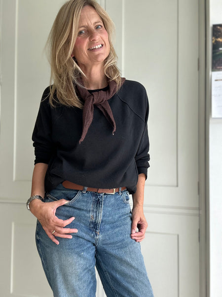 Woman wearing a black top and blue jeans with a brown scarf, standing against a white paneled wall.