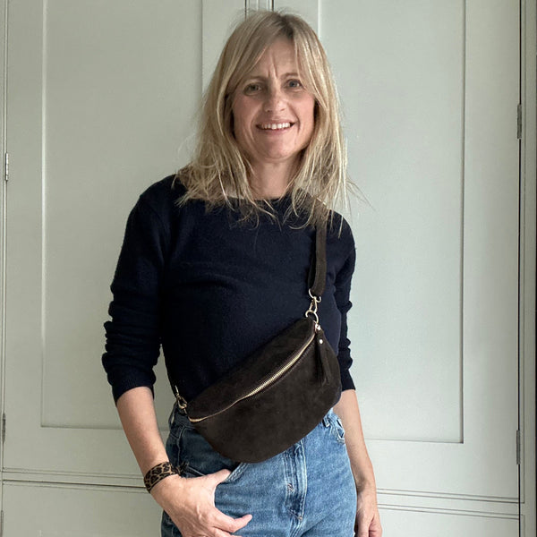 Woman wearing a black top and blue jeans with a brown bag, standing in a room with white walls.
