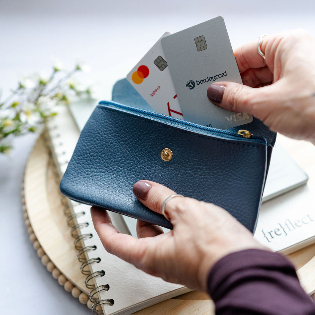 Person holding a blue wallet with cards, on a light surface with a notebook and plant.