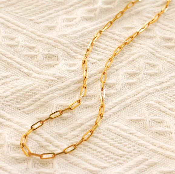 A gold paperclip chain necklace laid out on a textured cream knit fabric background.