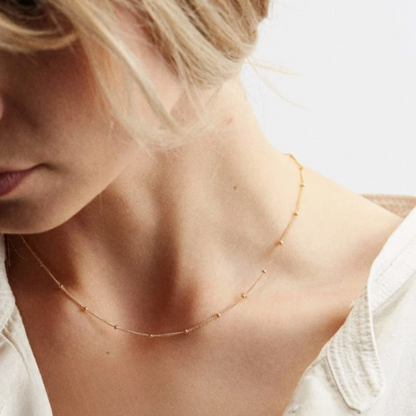 A close-up lifestyle shot of a model wearing a delicate gold chain necklace with small spaced beads, styled with a white top against a clean white background.