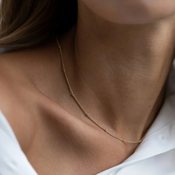 A close-up of a delicate gold chain necklace worn on a person's neck, styled with a white collared shirt.
