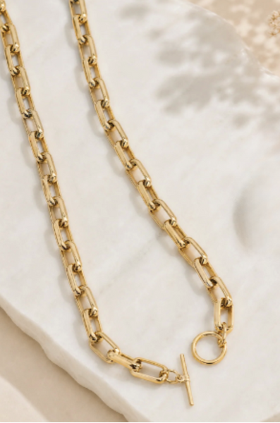 A gold-toned chain necklace with a toggle clasp and small circular charm, styled on a white stone slab with soft beige background shadows.